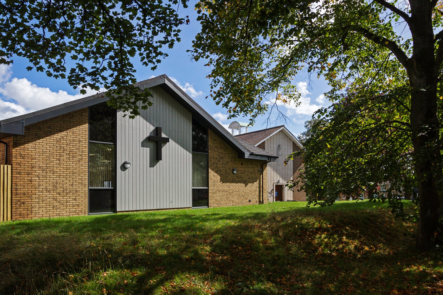 Croft Architecure Exterior view of a modern church building with a pitched roof, brick walls, and a vertical panel featuring a cross-shaped design, surrounded by green grass and mature trees under a partly cloudy sky.