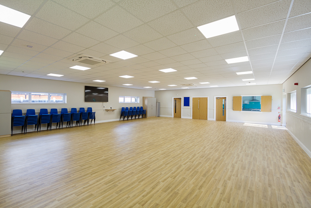 Croft Architecture Interior view of a spacious, modern hall with light wood flooring, white walls, and a suspended ceiling with recessed lighting. The room features stacked blue chairs along one wall, multiple doors, a wall-mounted screen, and several windows allowing natural light. Croft Architecture Interior view of a spacious, modern hall with light wood flooring, white walls, and a suspended ceiling with recessed lighting. The room features stacked blue chairs along one wall, multiple doors, a wall-mounted screen, and several windows allowing natural light.