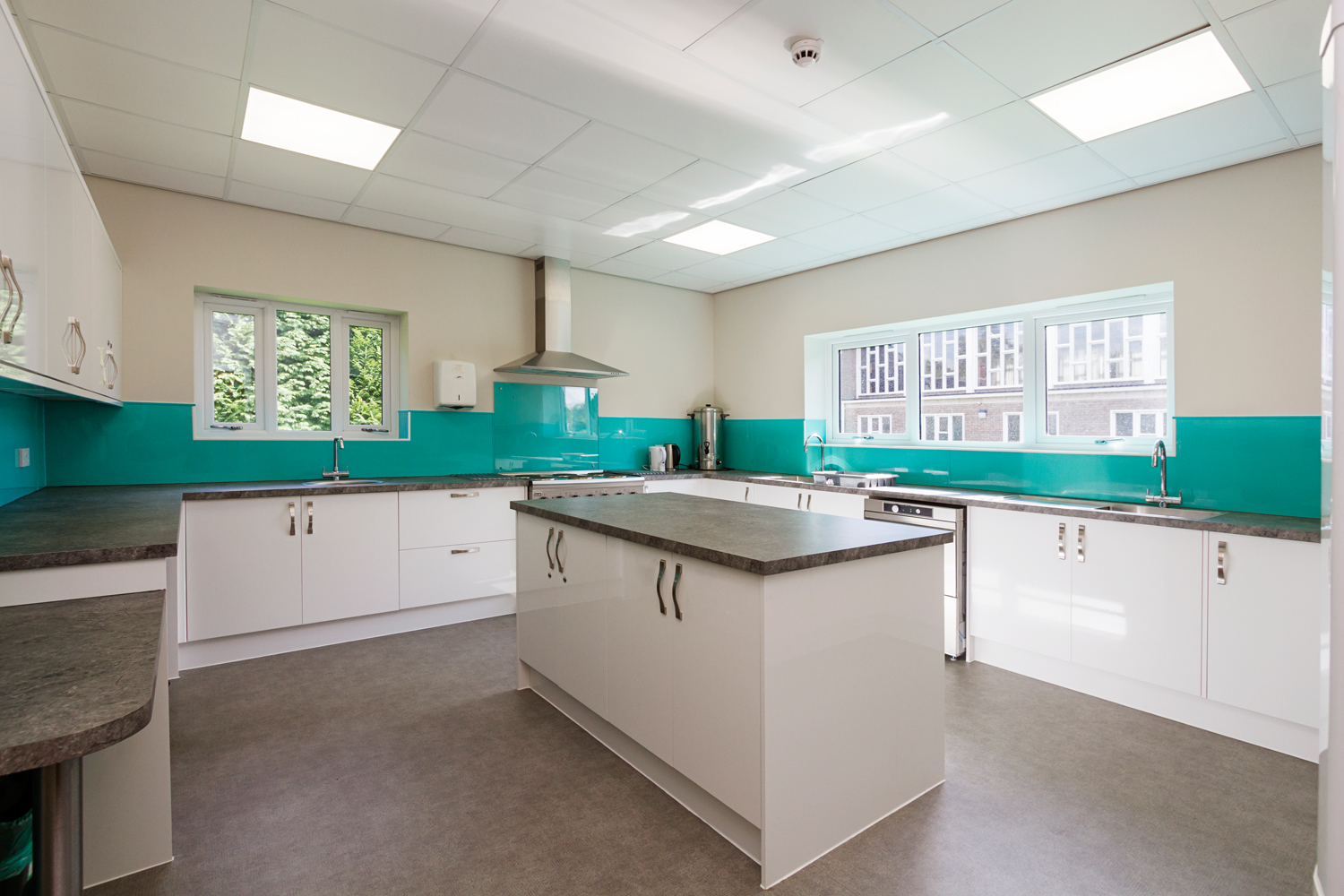 Modern kitchen interior designed by Croft Architecture, featuring white cabinetry, a central island with dark worktops, and vibrant turquoise splashbacks. Large windows provide natural light, creating a bright, functional space for community use. Modern kitchen interior designed by Croft Architecture, featuring white cabinetry, a central island with dark worktops, and vibrant turquoise splashbacks. Large windows provide natural light, creating a bright, functional space for community use.